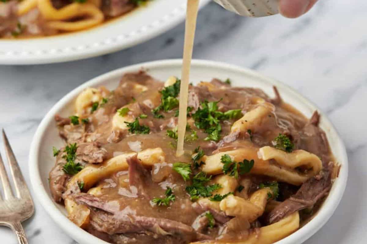 A white bowl filled with beef and noodles in a rich gravy, garnished with chopped parsley, is being drizzled with extra gravy from a small container. A silver fork is placed beside the bowl, and a second plate of the same dish can be seen in the background.