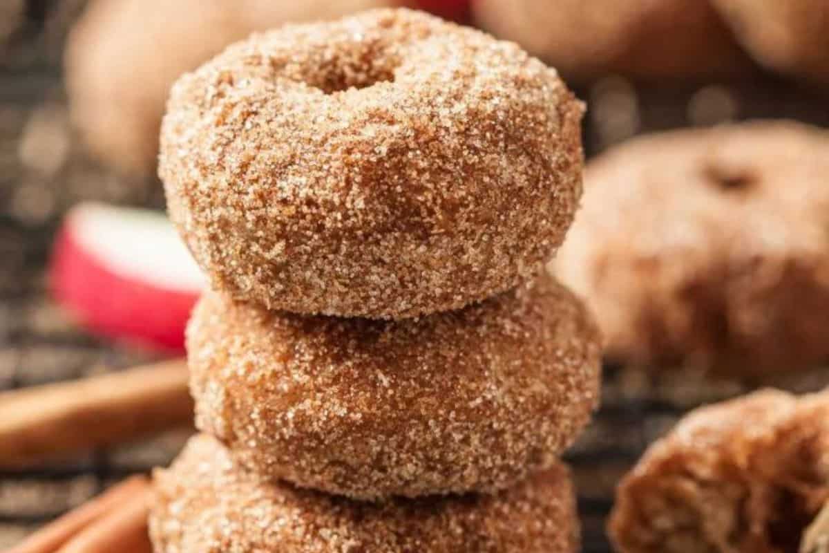 A close-up of a stack of three sugar-coated apple cider donuts. The donuts have a light brown hue and are generously coated with granulated sugar. The background shows additional donuts slightly out of focus, with a cinnamon stick and an apple slice visible.