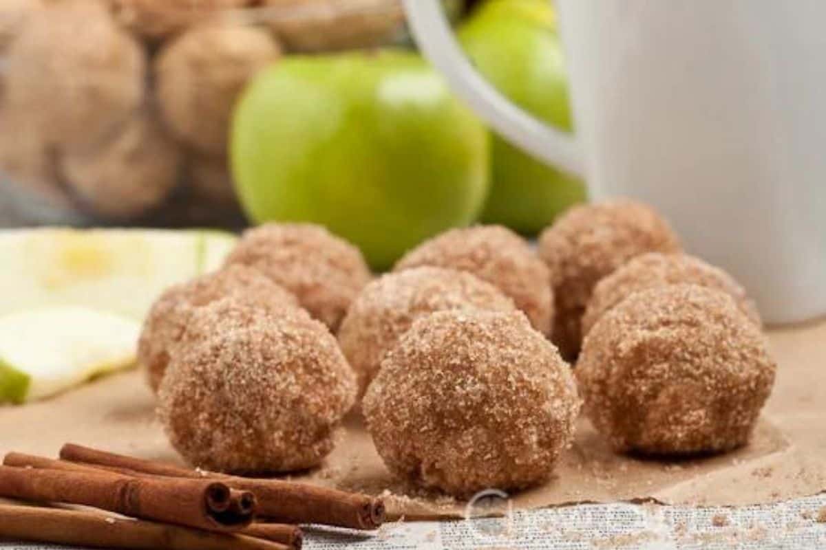 Close-up image of several apple cider donut holes coated in cinnamon sugar, arranged on a sheet of brown parchment paper. A pair of cinnamon sticks, a fresh green apple, and a white mug are blurred in the background.