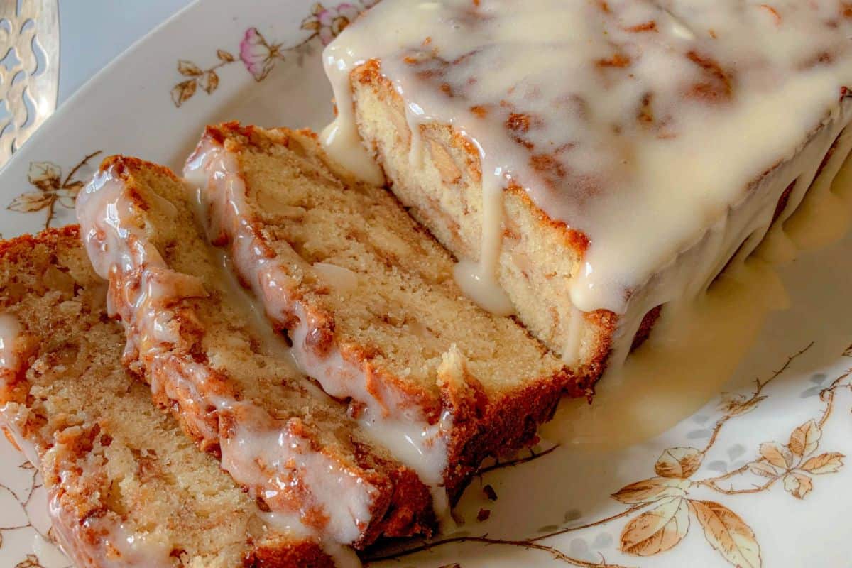 A close-up of a glazed loaf cake on a floral-patterned plate. The cake is partially sliced, showing a moist and dense interior. The icing generously drips over the top and down the sides, creating an inviting look.