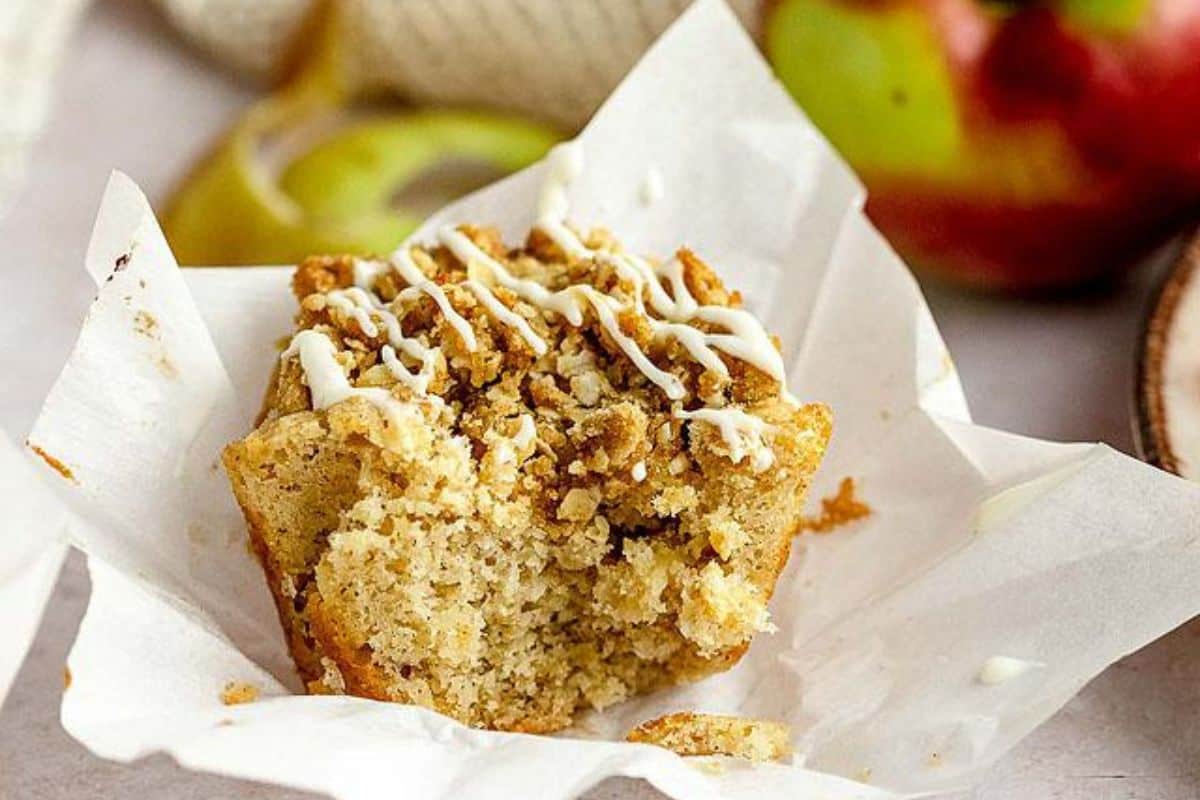 A partially eaten muffin with a crumbly top sits on white parchment paper. The muffin is topped with a white drizzle, likely icing. In the blurred background, there are apples and a coil of twine.