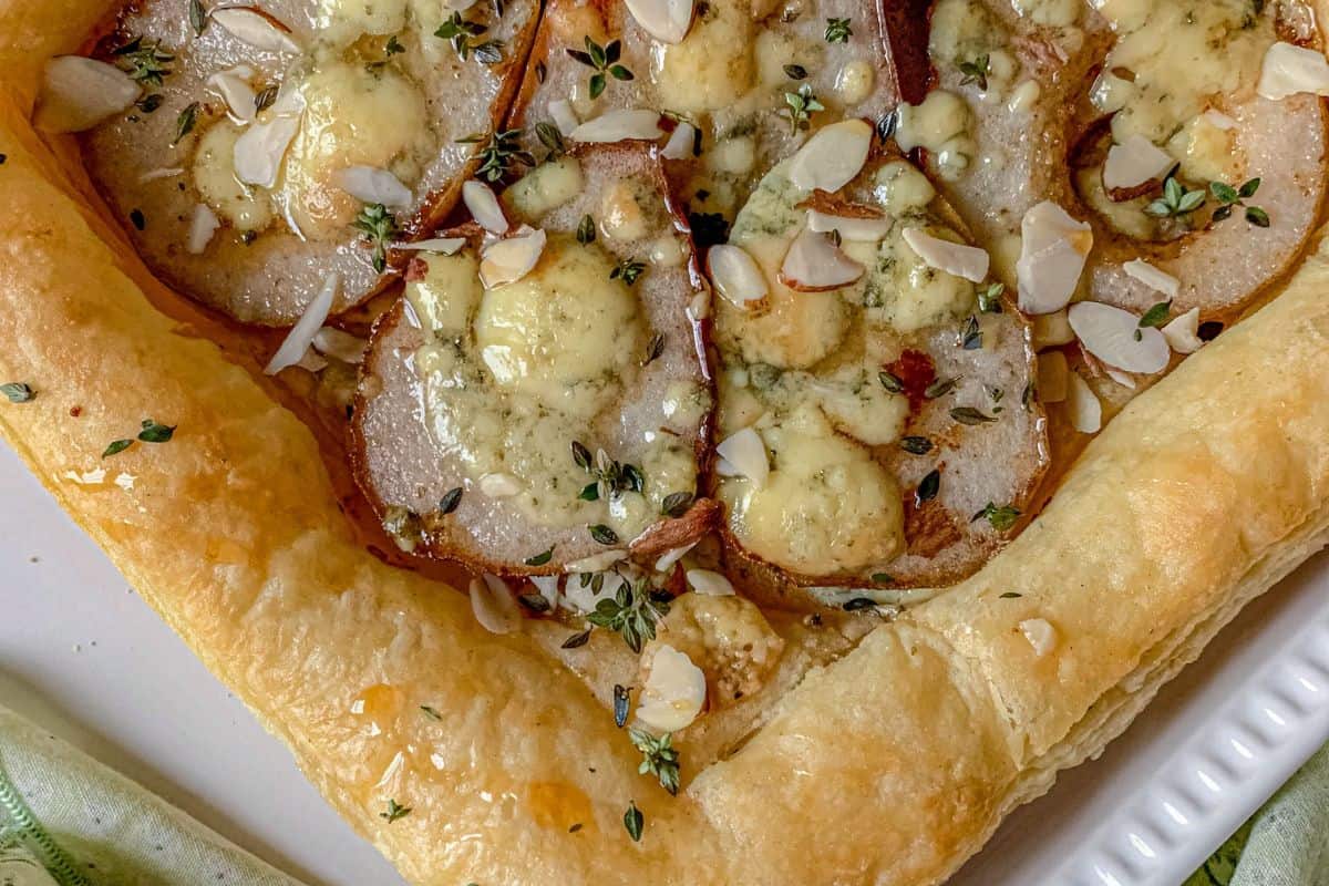 A close-up of a golden puff pastry topped with sliced pears, melted blue cheese, sliced almonds, and fresh thyme leaves.