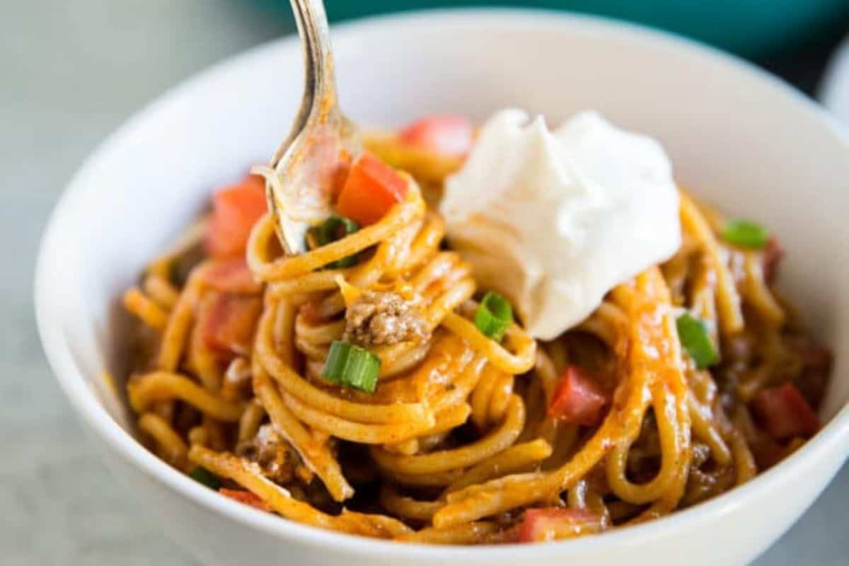A bowl of spaghetti topped with ground meat, diced tomatoes, sliced green onions, and a dollop of sour cream. A fork is twirling the spaghetti, lifting it from the bowl. The dish appears rich and creamy, with a hearty sauce coating the noodles.