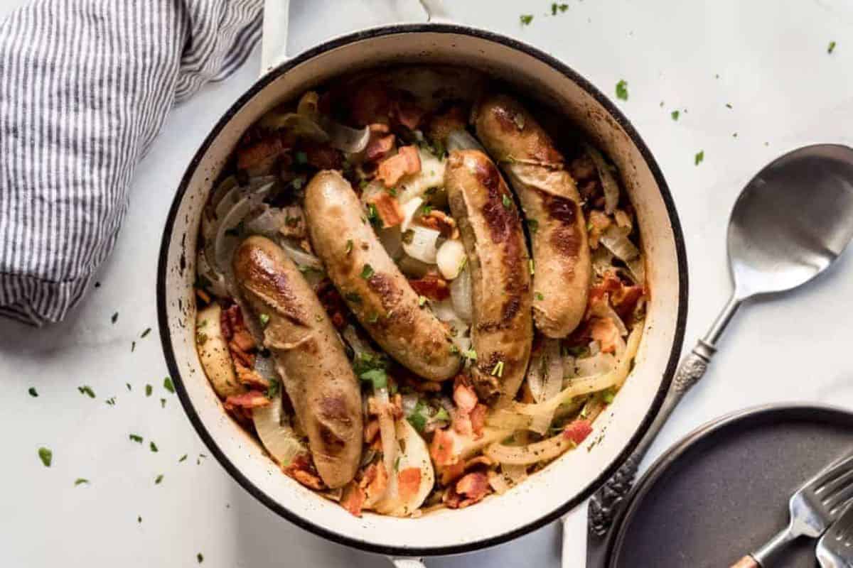 A round pot, partially filled with grilled sausages, sliced onions, and chopped bacon, garnished with herbs. The pot is on a white surface next to a striped cloth, a metal spoon, and a dark plate with a fork on it.