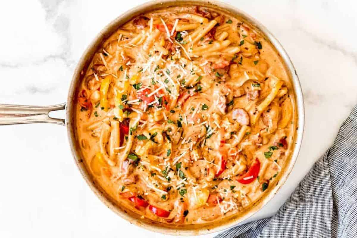 A skillet filled with creamy Cajun chicken pasta, featuring penne pasta, slices of chicken, colorful bell peppers, and a sprinkle of grated Parmesan cheese and chopped parsley on top. The skillet is placed on a white marble countertop next to a blue-striped towel.