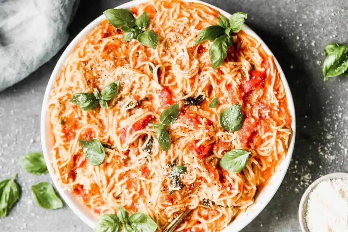 A bowl filled with spaghetti topped with a tomato-based sauce, fresh basil leaves, and grated cheese. The dish is set on a gray surface, with a few sprigs of basil scattered around the bowl.