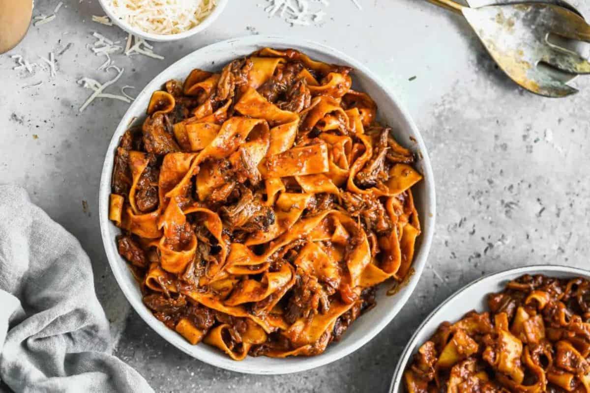 A white bowl filled with wide ribbons of pappardelle pasta coated in a rich, meaty ragu sauce, set on a gray surface. A smaller bowl of grated cheese and a cloth napkin are nearby, along with a serving spoon and fork.