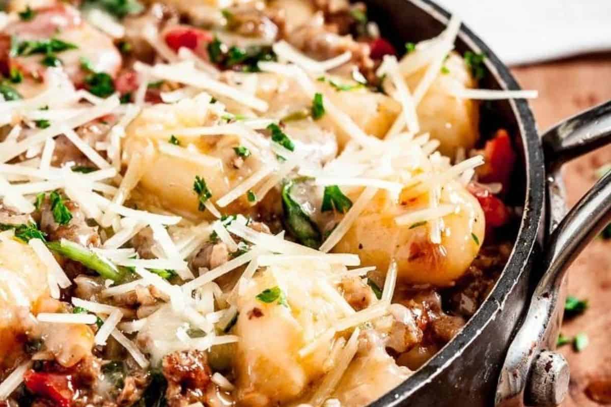 A close-up shot of a skillet filled with a gnocchi dish. The gnocchi is topped with ground meat, chopped tomatoes, spinach, melted cheese, and garnished with grated Parmesan and chopped parsley. The dish appears rich and savory with a variety of textures and colors.