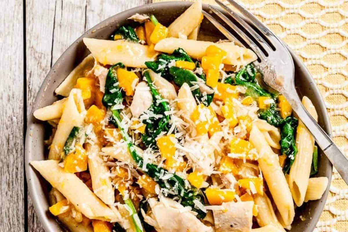 A close-up of a bowl of penne pasta mixed with vibrant yellow bell peppers, spinach leaves, and chunks of chicken. The dish is garnished with grated cheese, and a fork rests on the edge of the bowl. The background features a wooden surface and a yellow-patterned cloth.