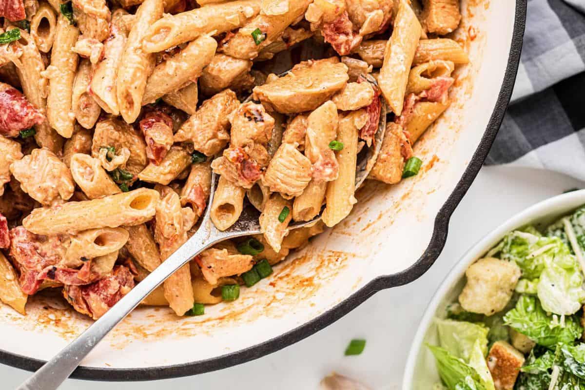 A close-up of a skillet filled with creamy penne pasta mixed with chunks of chicken and sun-dried tomatoes, garnished with chopped green onions. Next to the skillet is a bowl of Caesar salad with croutons. A checkered napkin is partially visible in the background.