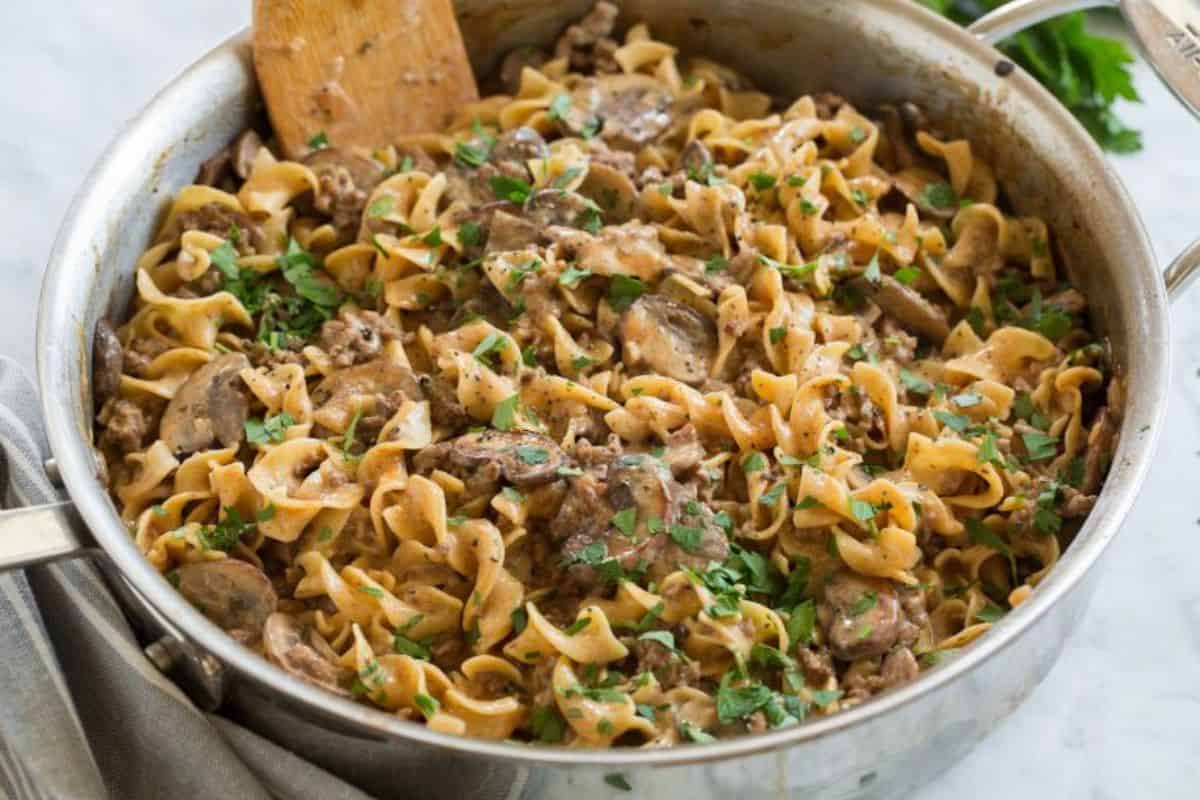 A large skillet contains a creamy beef stroganoff dish with wide egg noodles, sliced mushrooms, and chunks of beef. The dish is topped with chopped parsley and a wooden spatula rests inside the skillet.