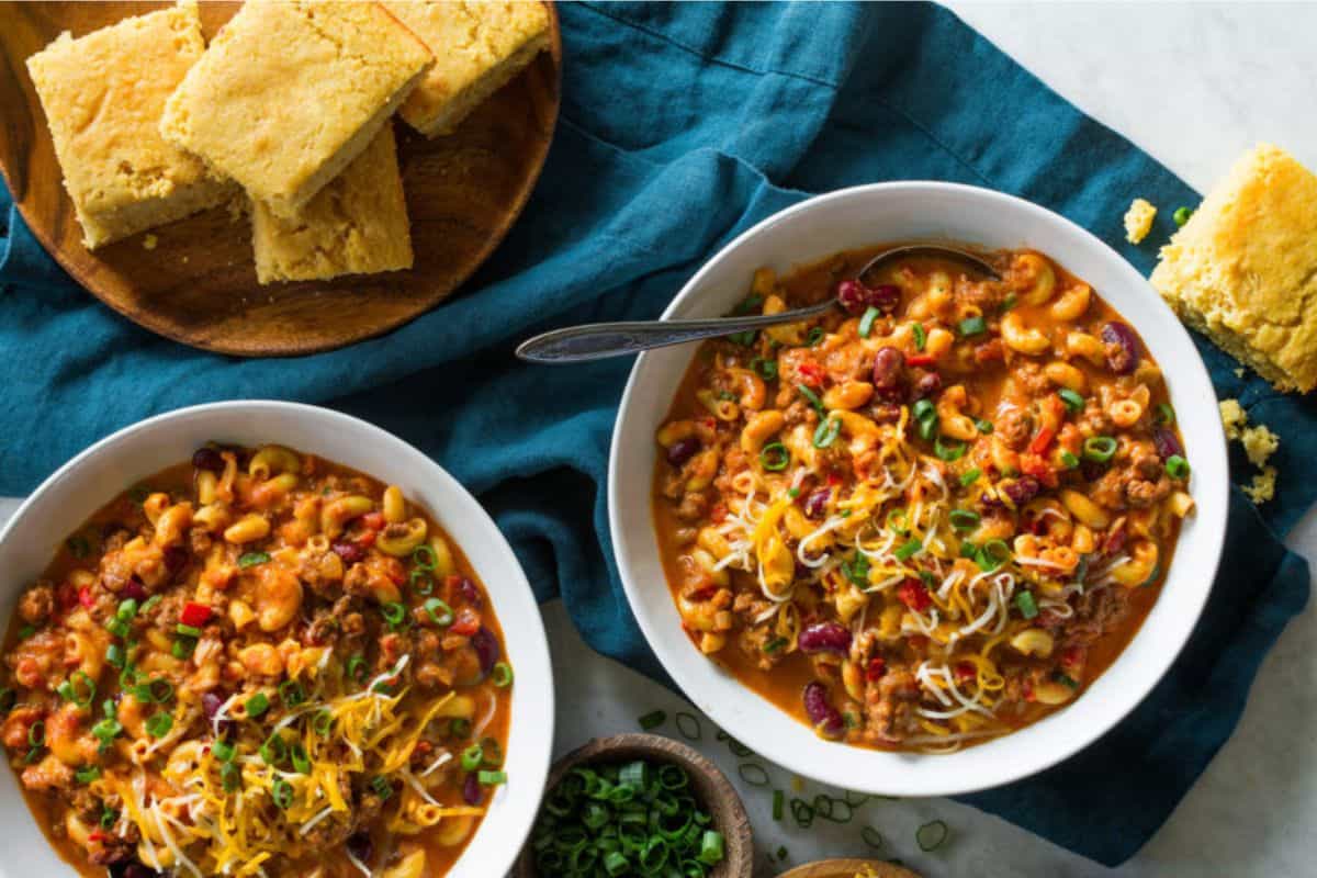 Two bowls of chili mac topped with shredded cheese and chopped green onions on a teal cloth. A spoon rests in one bowl. A wooden plate with cornbread slices and a few crumbs is placed to the side. A small bowl of chopped green onions is nearby.