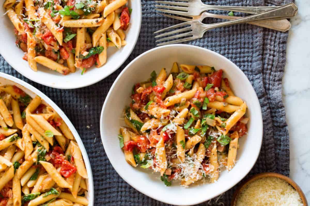 Three bowls of penne pasta topped with a tomato-based sauce, grated cheese, and fresh herbs are displayed on a gray textured cloth. Silver forks are placed beside the bowls, and a small wooden bowl of grated cheese is visible in the bottom right corner.