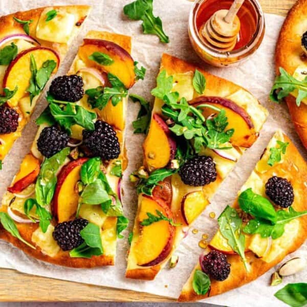 A flatbread pizza is topped with fresh blackberries, peach slices, arugula, red onion, and mozzarella cheese. The pizza is cut into four slices and garnished with pistachios. A small bowl of honey with a honey dipper is placed beside the pizza on a wooden board.