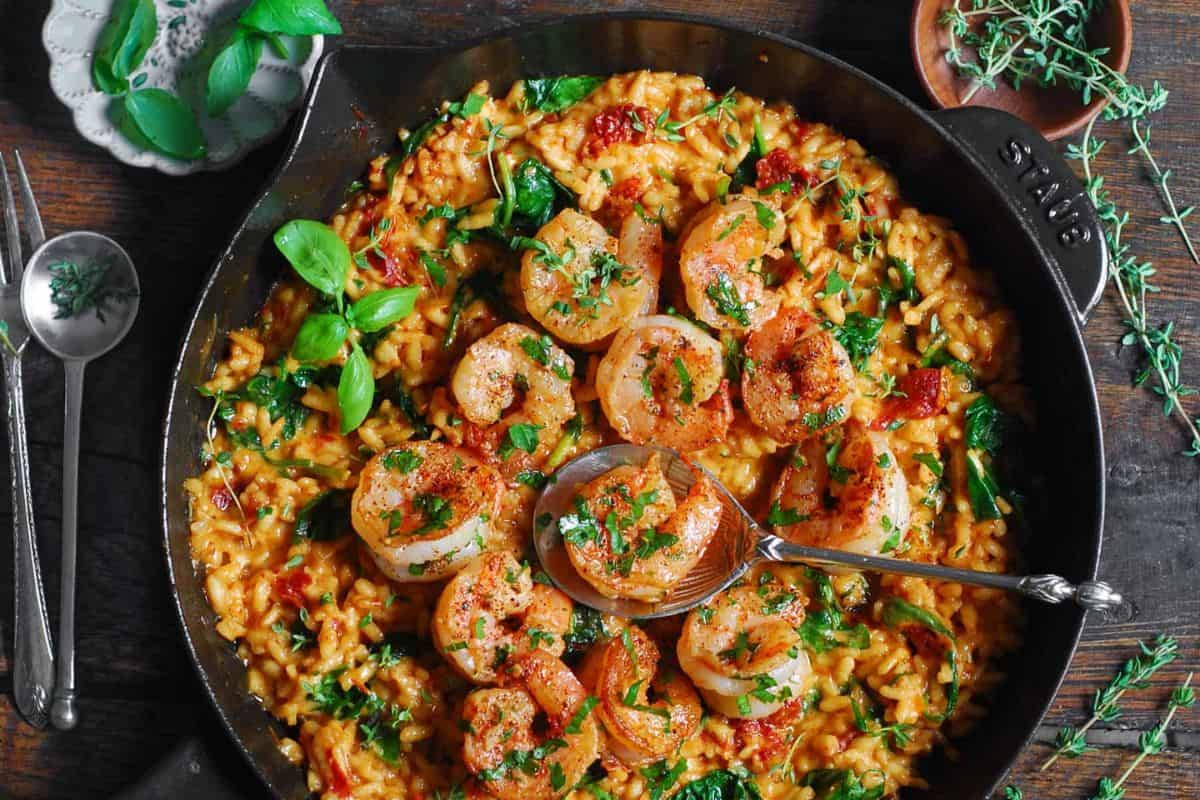 A skillet filled with creamy risotto topped with seared shrimp, garnished with fresh herbs. The dish contains pieces of sun-dried tomatoes and wilted spinach, with a spoon placed in the center. Fresh herbs are scattered around the skillet on the wooden table.