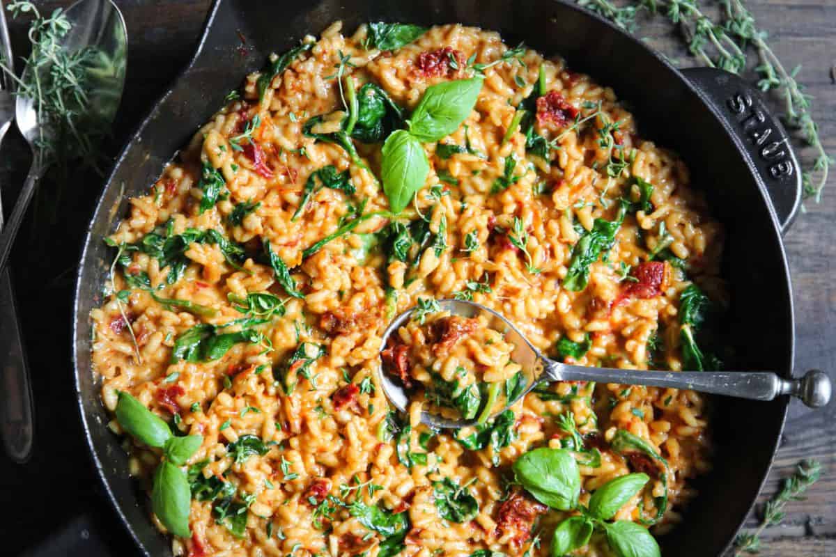 A skillet filled with creamy risotto mixed with sun-dried tomatoes, fresh spinach, and garnished with basil leaves. A large spoon is resting in the skillet, ready to serve. Fresh sprigs of herbs are scattered around the pan on a dark wooden surface.