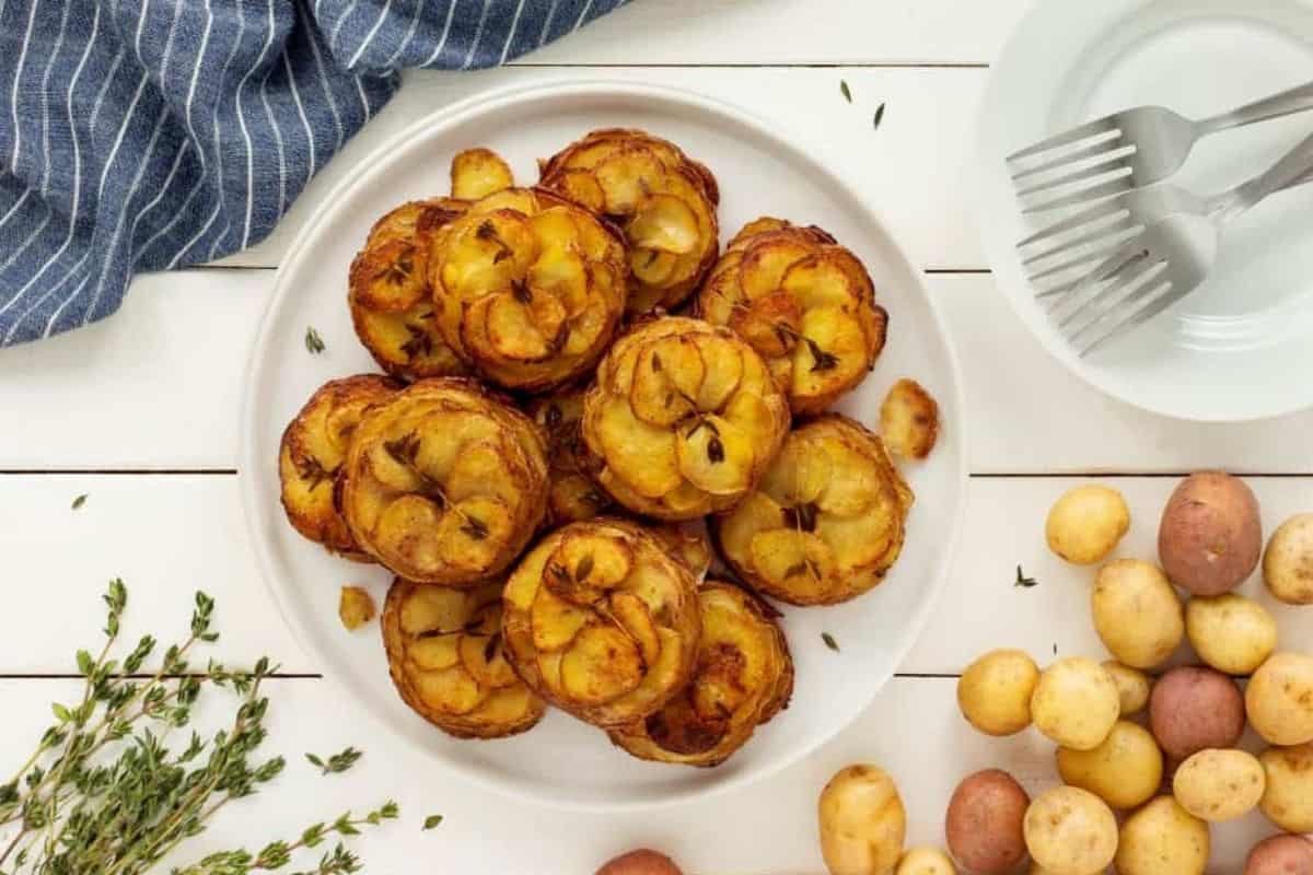 A white plate filled with golden-brown, cooked potato rounds garnished with fresh herbs. The plate is placed on a white wooden table with an herb sprig, a bunch of small potatoes, a small white bowl with forks, and a blue-striped cloth nearby.