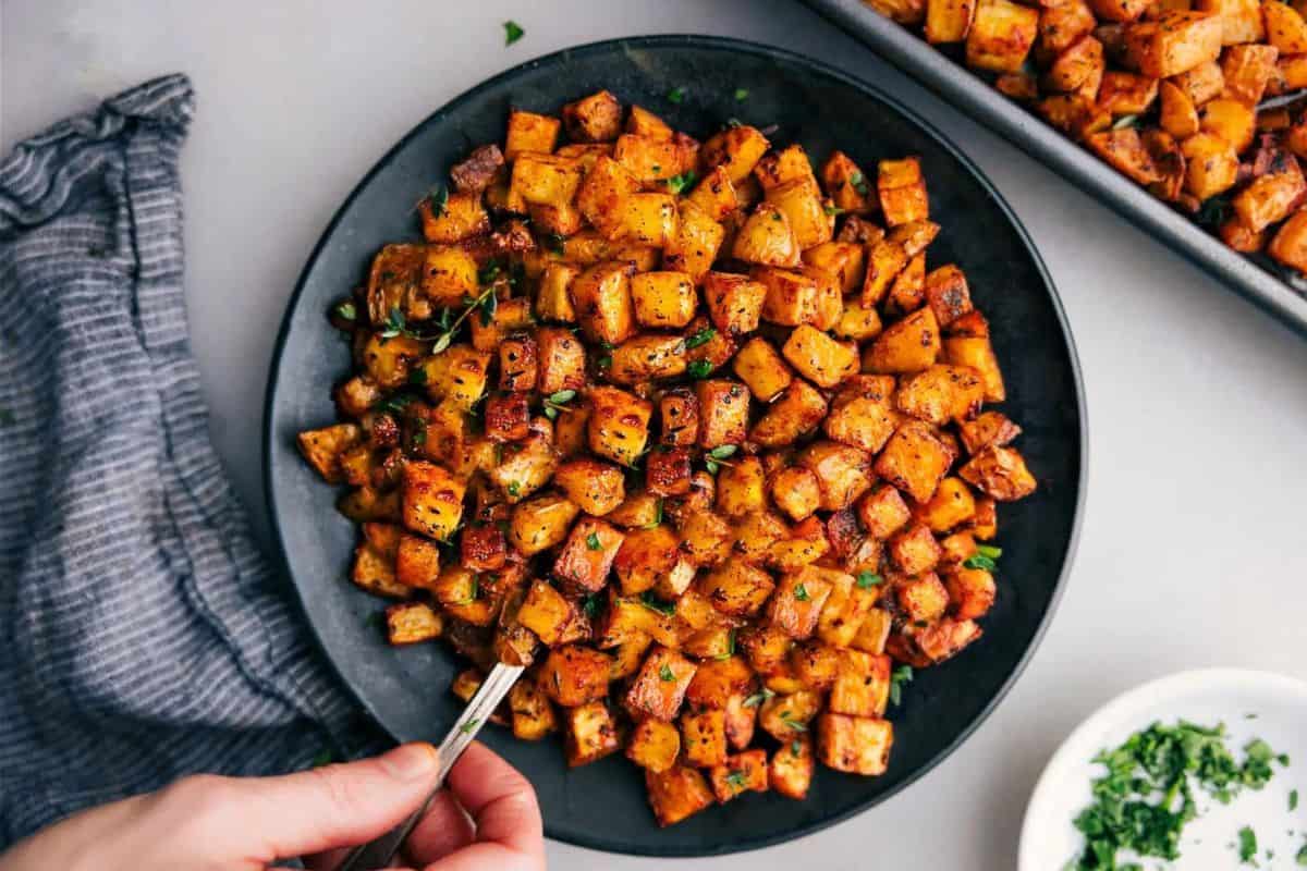 A top-down view of a black plate filled with golden-brown roasted potatoes garnished with herbs. A hand is seen holding a fork, ready to take a bite. A baking sheet with more roasted potatoes is visible in the background along with a small bowl of chopped herbs.