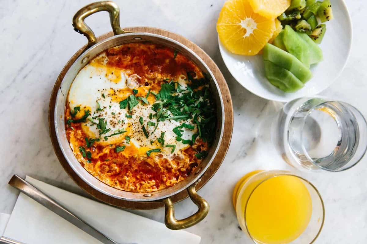 A bowl of Shakshuka garnished with herbs and placed on a marble surface next to a plate of sliced kiwi and orange. Also on the table are a glass of water, a glass of orange juice, and folded napkins with a fork placed on top.