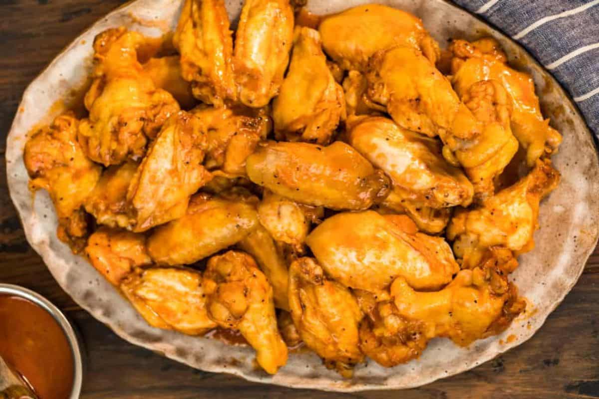 A large platter filled with golden-brown chicken wings, lightly coated in sauce. The wings are arranged in a rustic, oval-shaped ceramic dish. A small bowl of dipping sauce is partially visible in the bottom left corner, and a striped cloth is in the top right.
