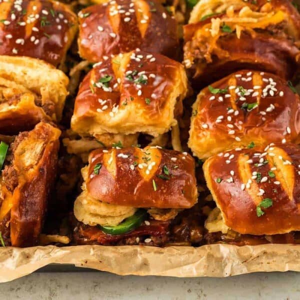 A tray filled with several freshly baked sliders. Each slider, made with soft pretzel buns, is stuffed with beef, melted cheese, jalapeño slices, and crispy fried onions. The buns are topped with sesame seeds and a sprinkle of chopped herbs.