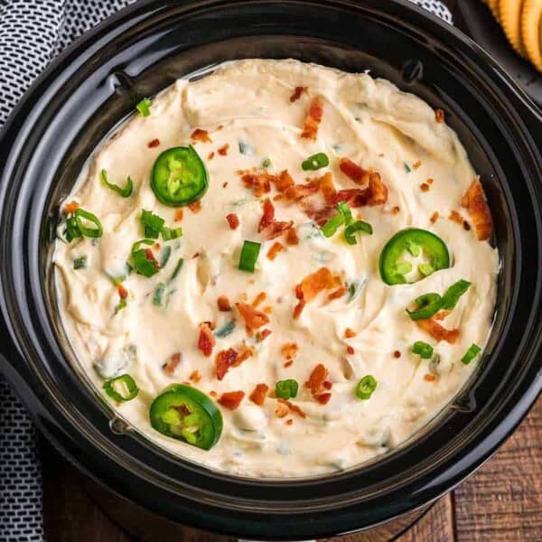 A slow cooker filled with creamy jalapeño dip, topped with sliced jalapeños, chopped green onions, and bacon bits. A plate of crackers sits in the background.