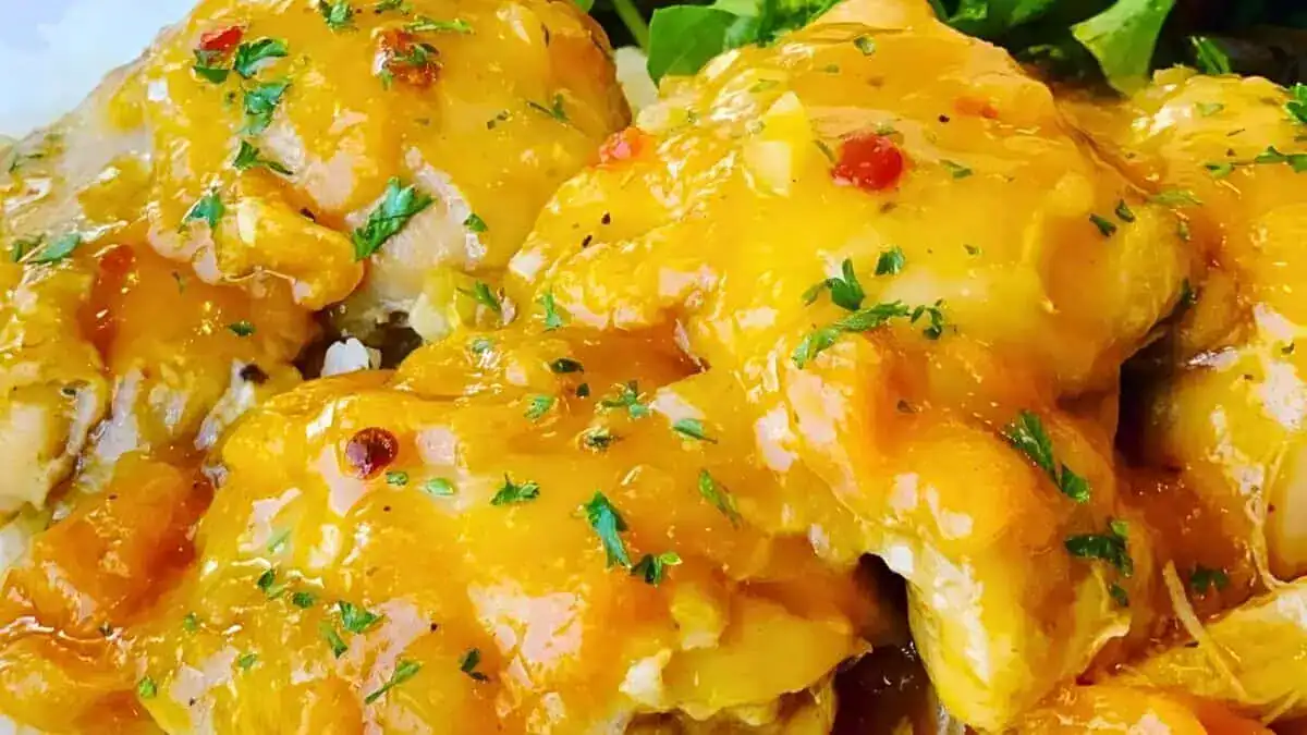 Close-up of three pieces of chicken covered in a bright, glossy, orange sauce garnished with chopped parsley. The chicken is served with greens in the background. The dish appears savory and well-prepared.