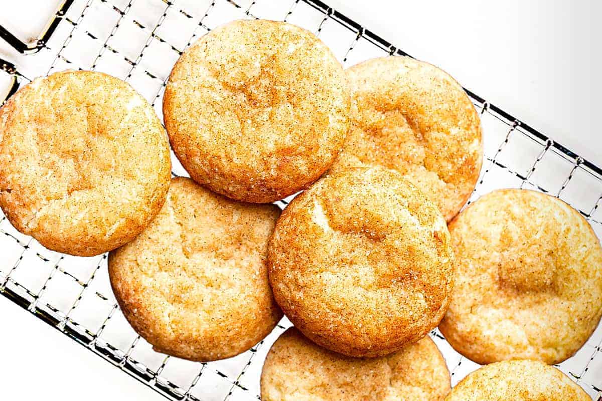 A batch of golden-brown snickerdoodle cookies resting on a cooling rack. The cookies have a slightly cracked surface, dusted with a light coating of cinnamon and sugar, highlighting their soft and chewy texture.