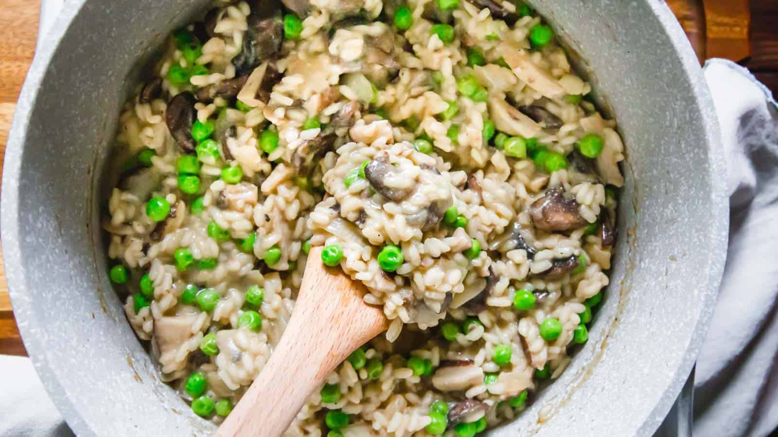 A creamy mushroom risotto with green peas is being stirred in a pot with a wooden spoon. The risotto is rich and has visible chunks of mushrooms and vibrant green peas scattered throughout the dish. The pot rests on a white cloth.
