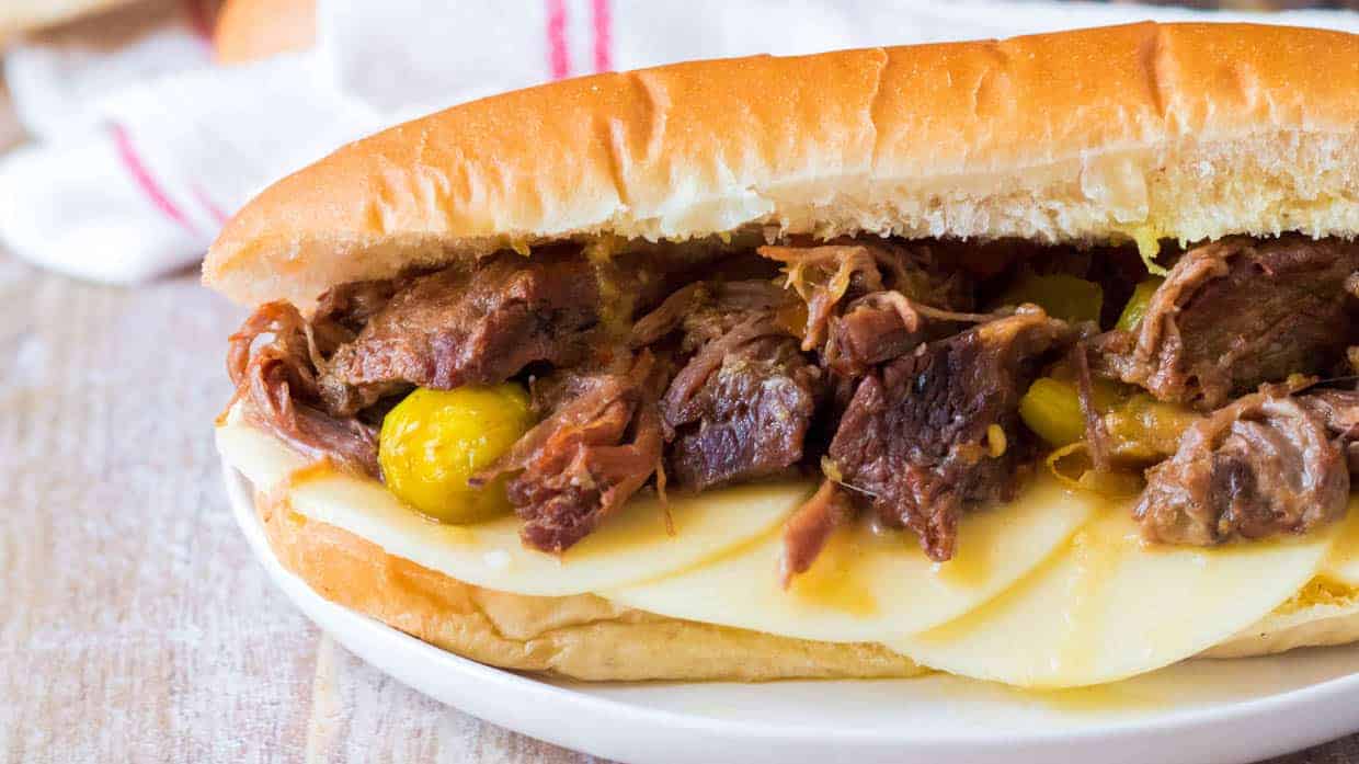 A close-up of a sandwich on a white plate. The sandwich is filled with tender, shredded meat, pickled vegetables, and topped with slices of cheese on a soft hoagie roll. A blurred cloth is visible in the background.