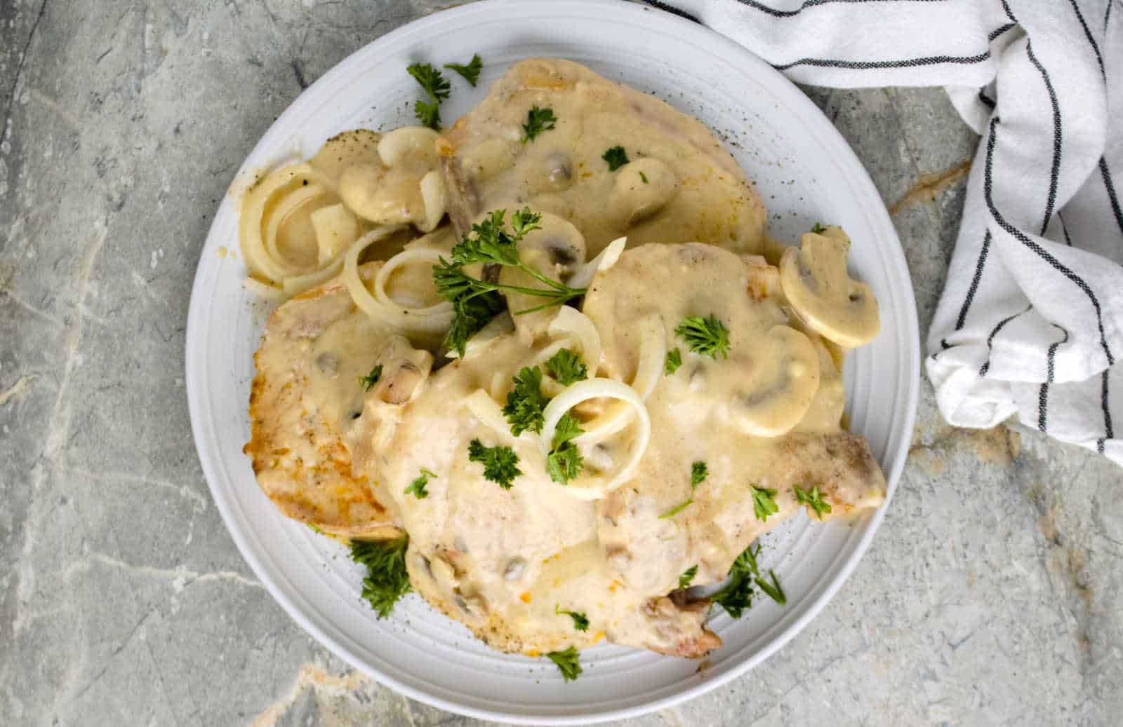 A white plate holds pork chops covered in a creamy mushroom and onion sauce, garnished with fresh parsley. A white and black striped cloth napkin is partially visible to the side on a rustic, textured surface.