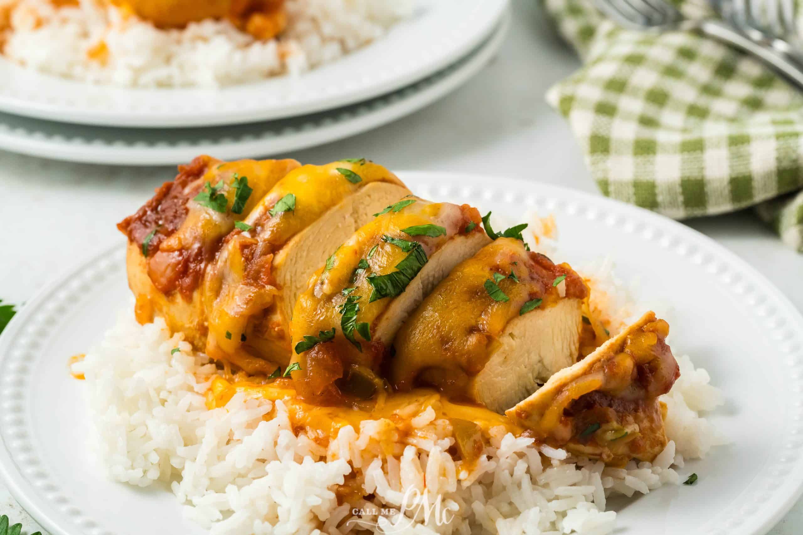 A plated dish features a baked chicken breast, sliced and topped with melted cheese and herbs, served on a bed of white rice. In the background, another plate is partially visible, along with a green checkered napkin and silverware.