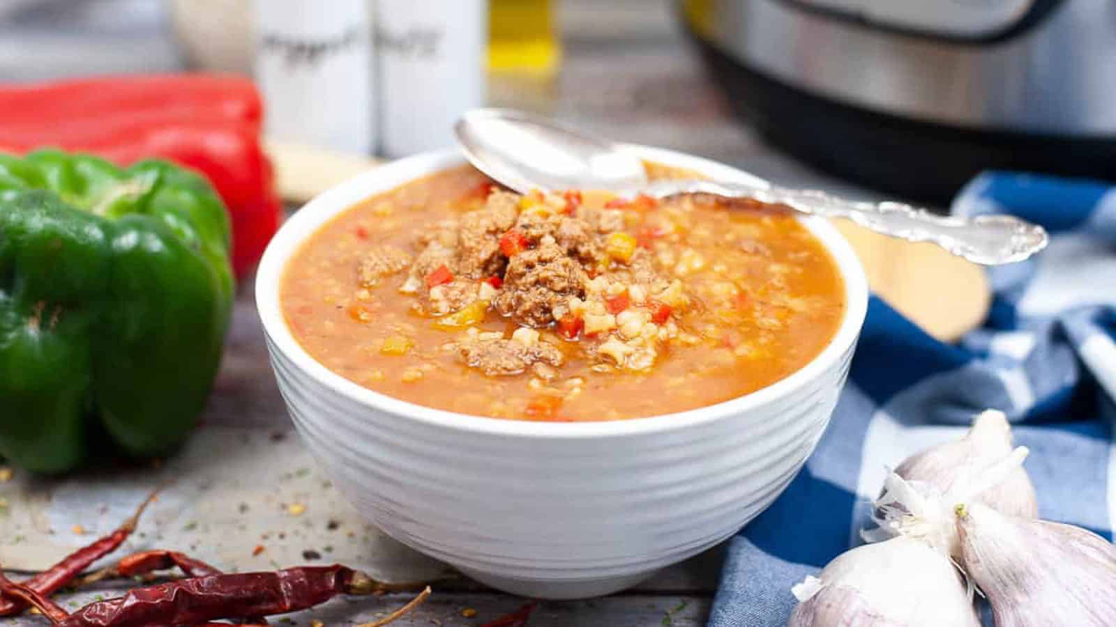 A bowl of hearty soup sits on a table, filled with ground meat, diced vegetables, and rice. A spoon rests on the rim of the white bowl. Surrounding the bowl are a green bell pepper, a red bell pepper, dried chili peppers, and some garlic cloves.