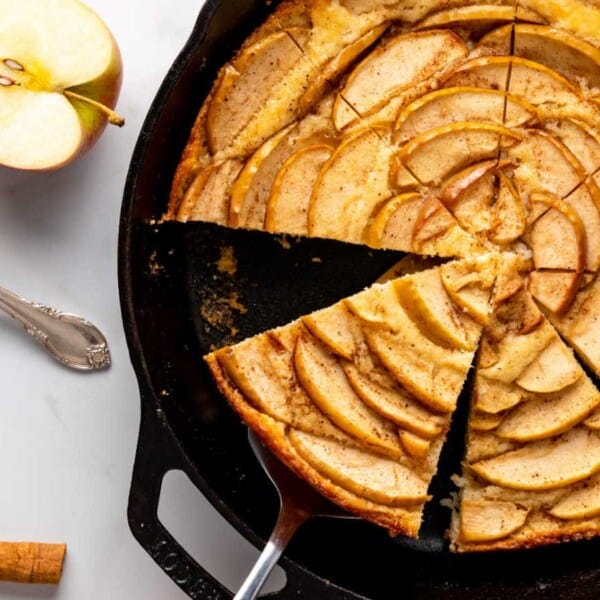 A sliced apple tart in a cast iron skillet with a server lifting one piece. Nearby are a plate, fork, tart slice, and half an apple.