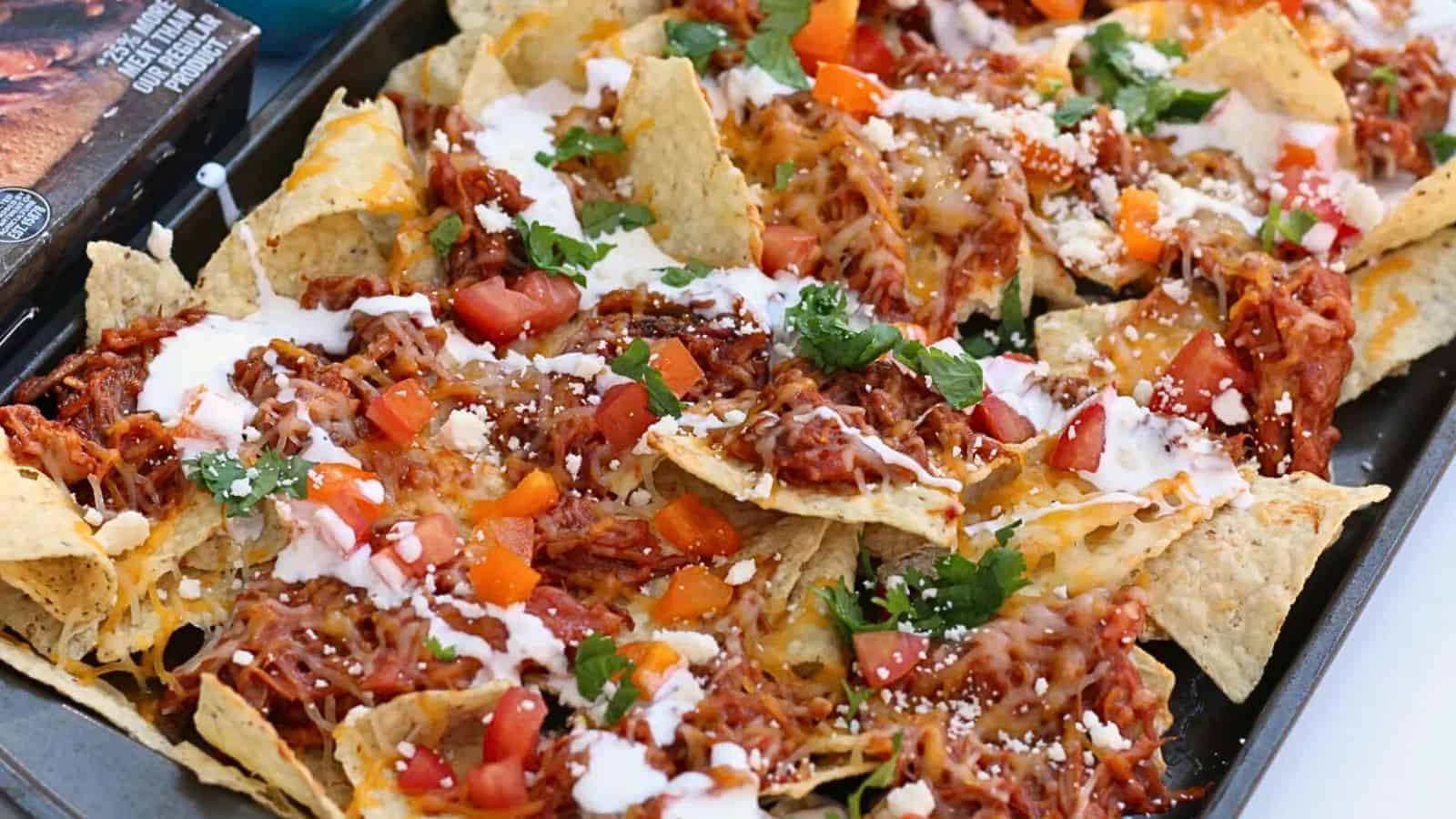 A tray of loaded nachos topped with shredded cheese, diced tomatoes, shredded meat, and drizzled with sour cream. Fresh cilantro is sprinkled on top for garnish. The nachos rest on a metal baking sheet.