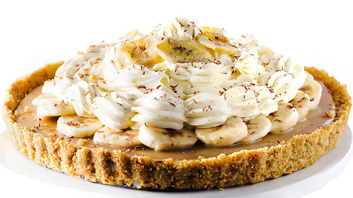 A banoffee pie on a white background. The pie has a crumbly golden crust, a layer of caramel, and is topped with sliced bananas and generous swirls of whipped cream, garnished with chocolate shavings.