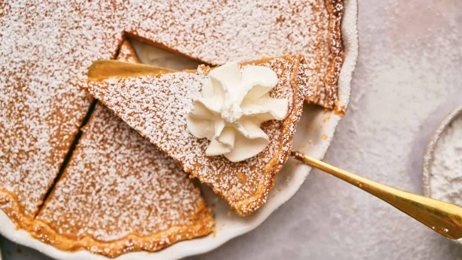 A close-up of a pumpkin pie with one slice being lifted out on a golden pie server. The pie is dusted with powdered sugar, and the lifted slice is topped with a dollop of whipped cream. The background is a light-colored surface.