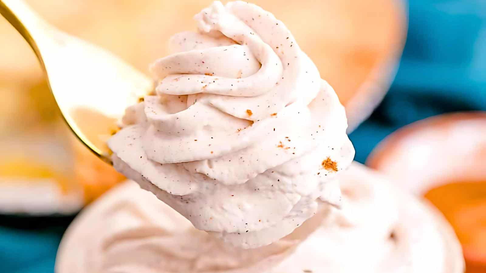 A close-up of a spoon holding a swirl of fluffy, creamy white dessert with specks of spice. The background is blurred, featuring hints of blue and orange, possibly indicating plates or additional servings of the dessert.