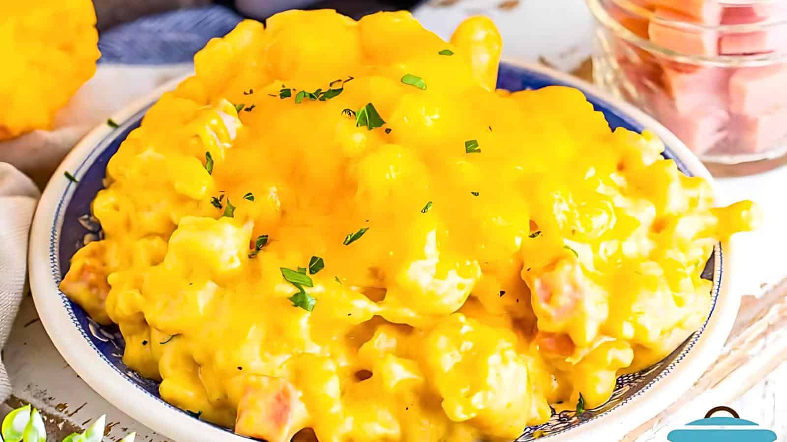 A blue-rimmed bowl of creamy mac and cheese topped with melted cheddar and garnished with chopped herbs. The dish is placed on a white wooden surface, surrounded by various small ingredients and utensils.