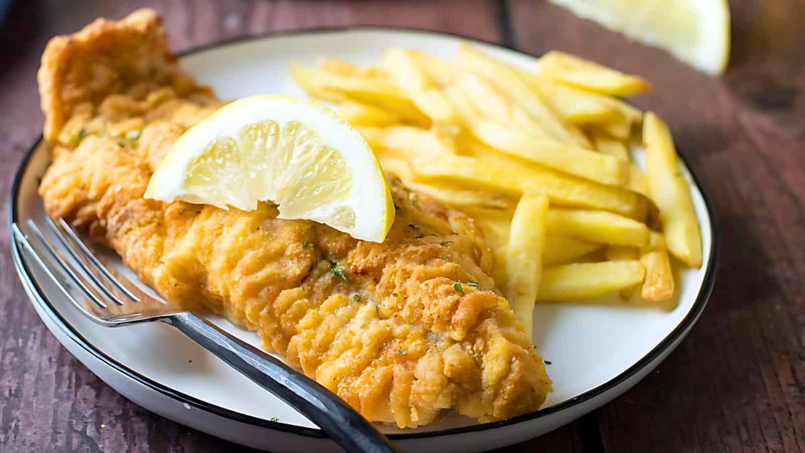 A plated serving of golden crispy fried fish with a wedge of lemon on top, accompanied by a side of French fries. A fork rests on the plate beside the food. The meal is presented on a white plate with a dark rim, set on a wooden table.