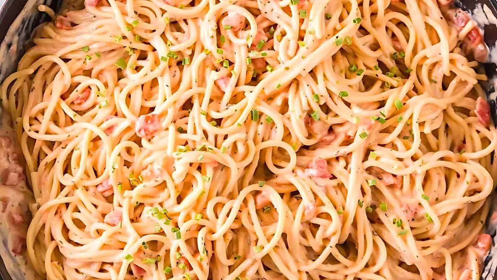 A close-up image of creamy spaghetti carbonara in a pan. The dish features a generous amount of spaghetti noodles mixed with bits of pancetta, coated in a creamy sauce, and garnished with finely chopped chives.