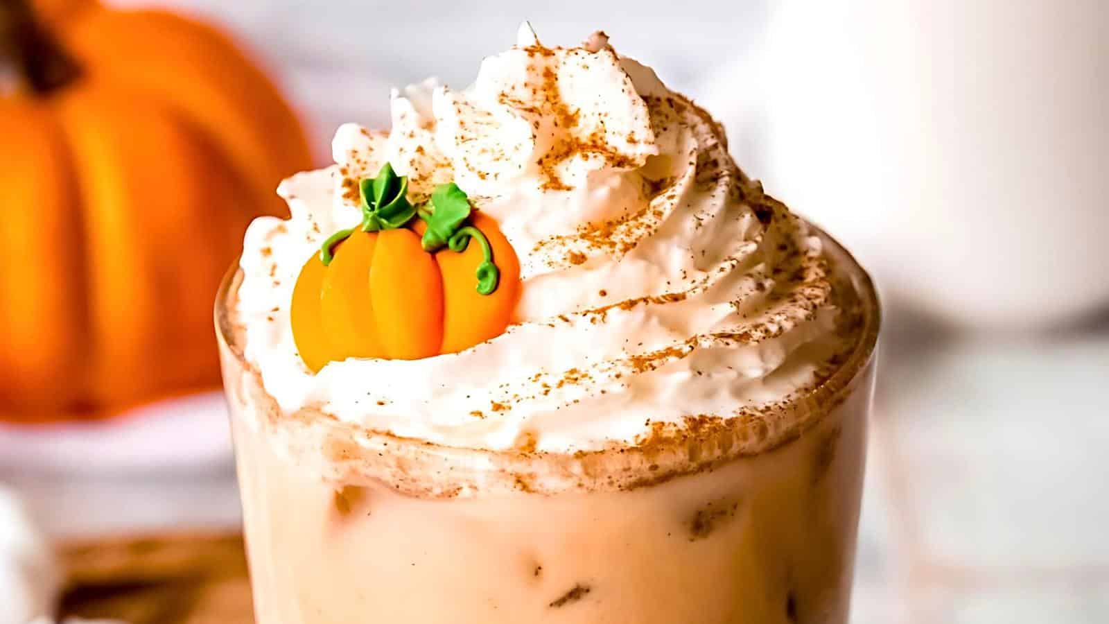 A close-up of a pumpkin spice latte topped with whipped cream and a small, decorative pumpkin candy. The drink is served in a clear glass with ice, with a blurred-out pumpkin in the background, evoking a cozy autumn vibe.