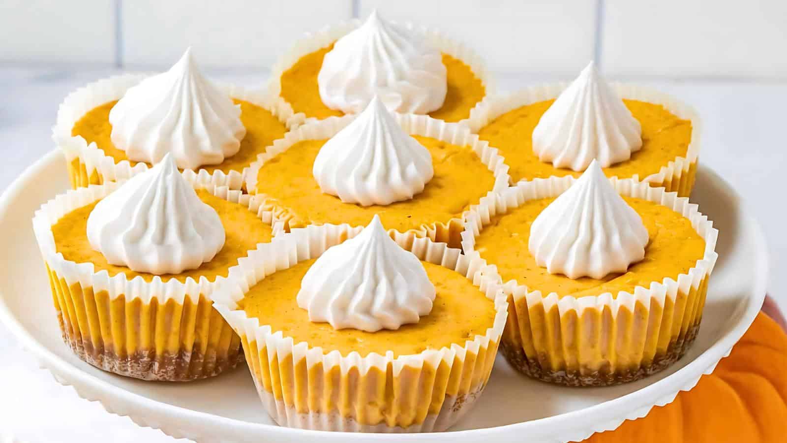 A plate of six mini pumpkin cheesecakes each topped with a swirl of whipped cream. The cheesecakes are in white cupcake liners and have a crumbly brown crust at the bottom. The background is a light-colored kitchen countertop.