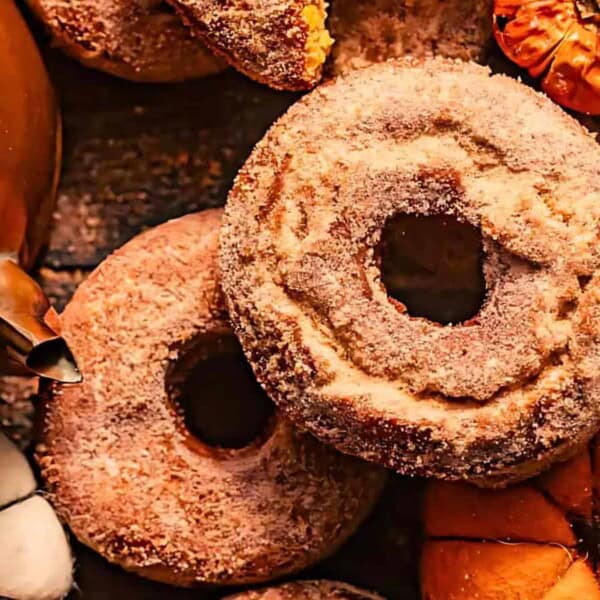 A close-up of sugared donuts surrounded by decorative pumpkins and part of a copper teapot on a rustic wooden surface.