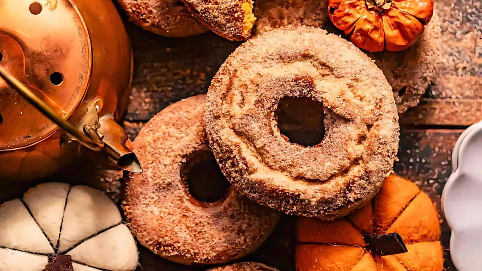 An assortment of sugar-coated donuts is arranged with decorative mini pumpkins on a rustic wooden surface. A copper teapot is positioned to the left, adding a touch of warmth to the autumnal scene.