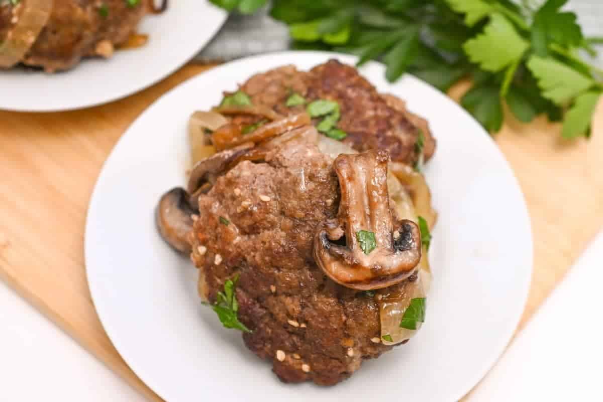 A plate of homemade Salisbury steak featuring two well-cooked beef patties garnished with sautéed mushrooms and onions. Fresh parsley is sprinkled on top, adding a touch of green. The plate sits on a wooden surface with a bunch of fresh parsley in the background.