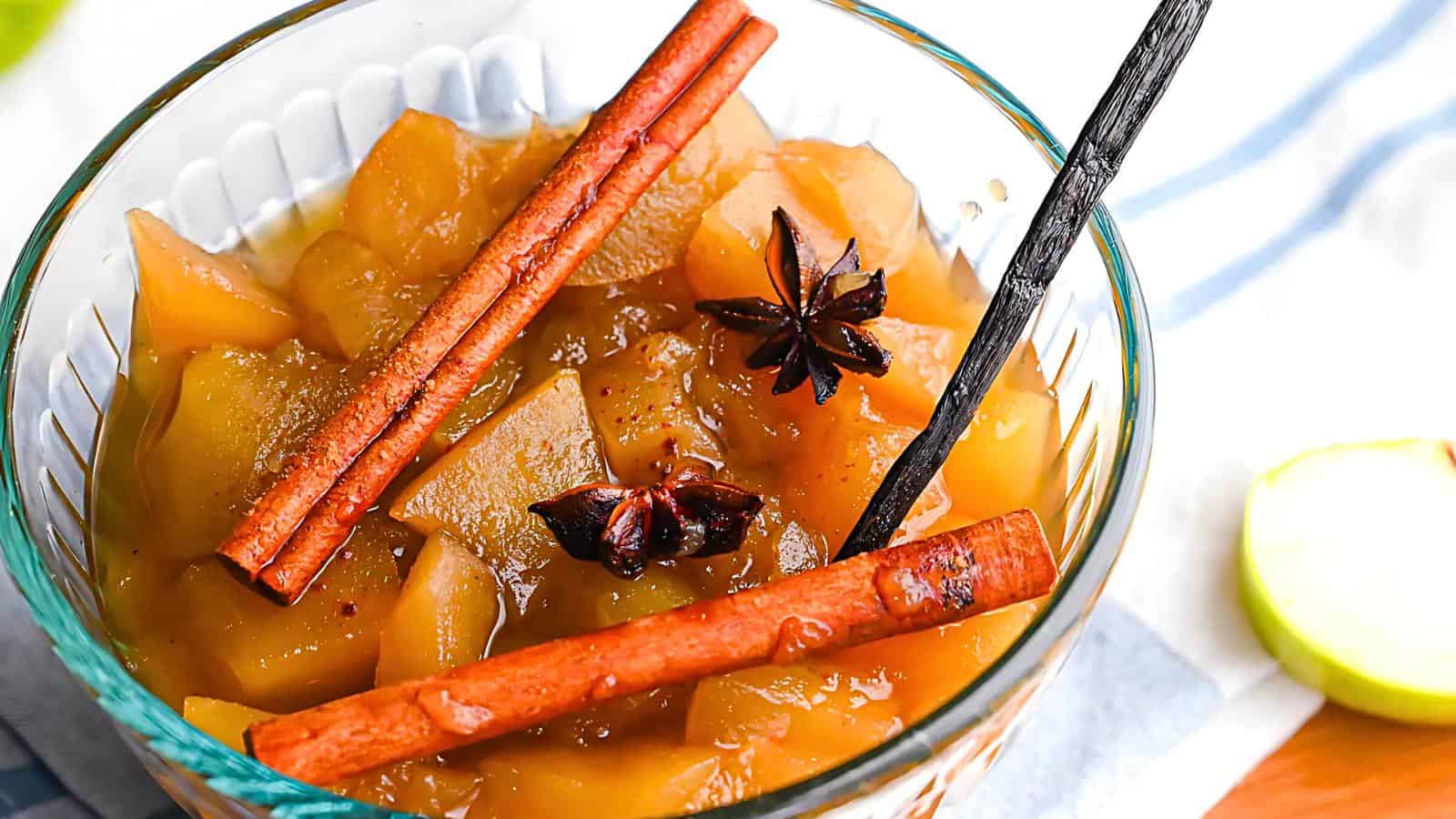 A glass bowl filled with a sweet apple compote, garnished with whole cinnamon sticks and star anise. The compote consists of chunky, glazed apple pieces. A wooden spoon rests inside the bowl. A green apple and a cloth napkin are partially visible in the background.