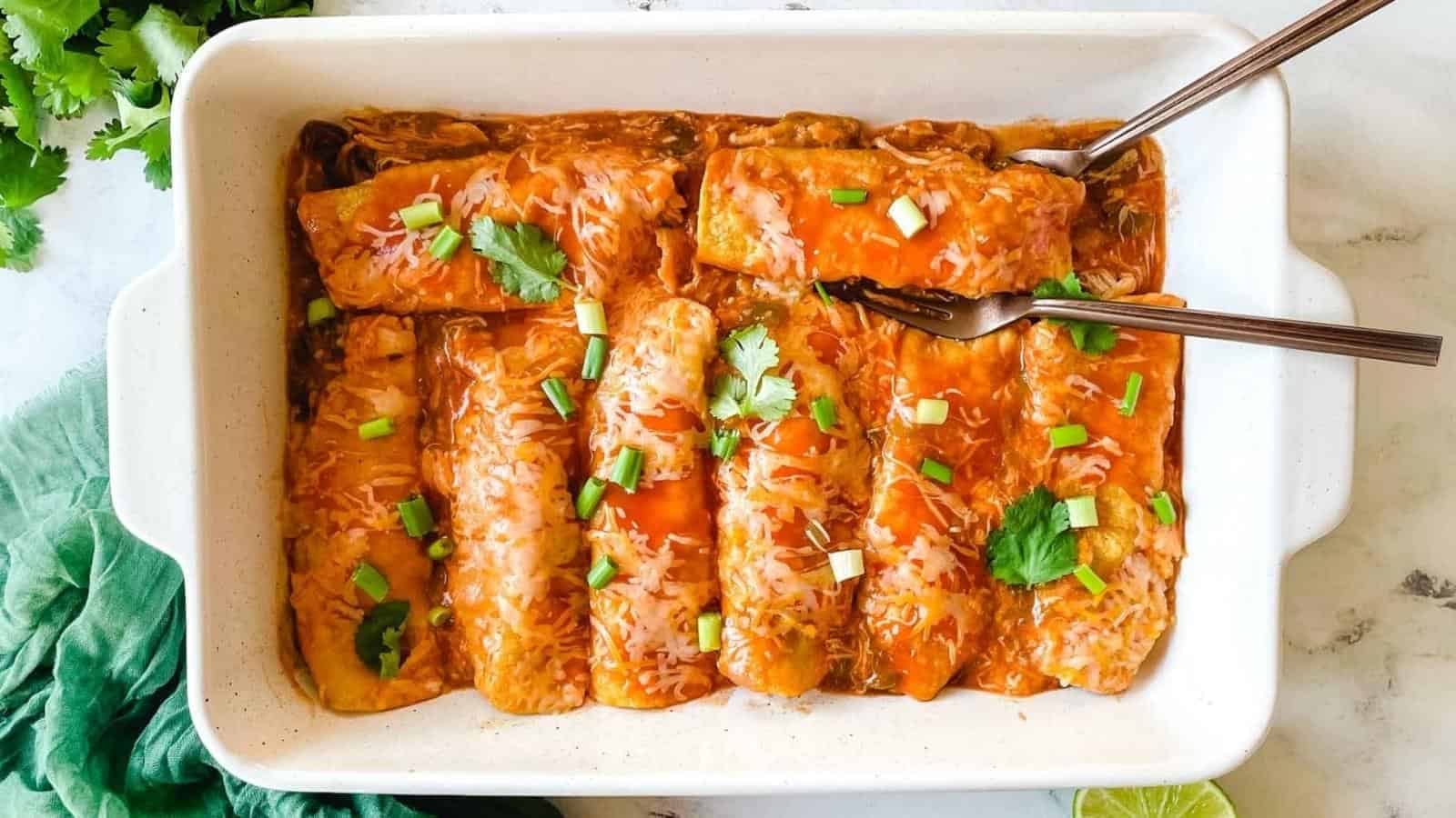 A rectangular white dish contains six enchiladas topped with melted cheese, red sauce, and garnished with chopped green onions and cilantro. Two serving utensils are placed in the dish, one on the left side and the other on the right side, ready to serve.