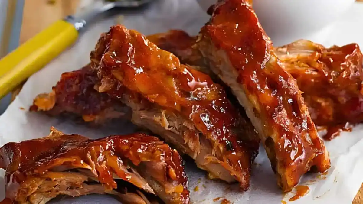 Close-up of a plate of glazed barbecue ribs. The ribs are covered in a rich, glossy sauce and are arranged on a piece of parchment paper. A yellow-handled utensil and a white bowl are in the blurred background. The ribs appear tender and juicy.