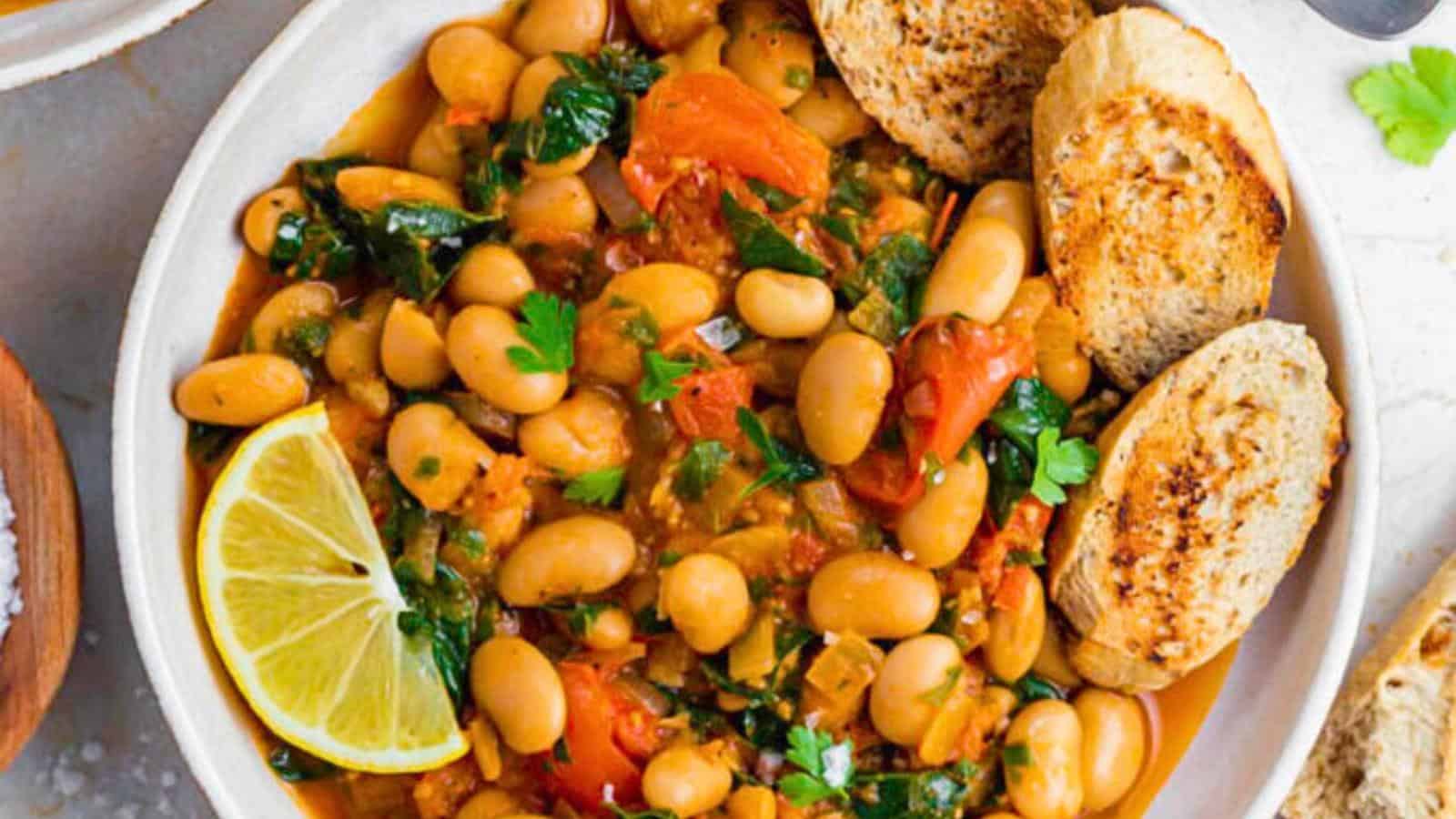 A vibrant bowl of mixed bean stew with tomatoes, spinach, and chopped herbs, garnished with a lemon wedge. The stew is served with two slices of toasted bread on the side. Fresh herbs add a touch of color on the edge of the white bowl.